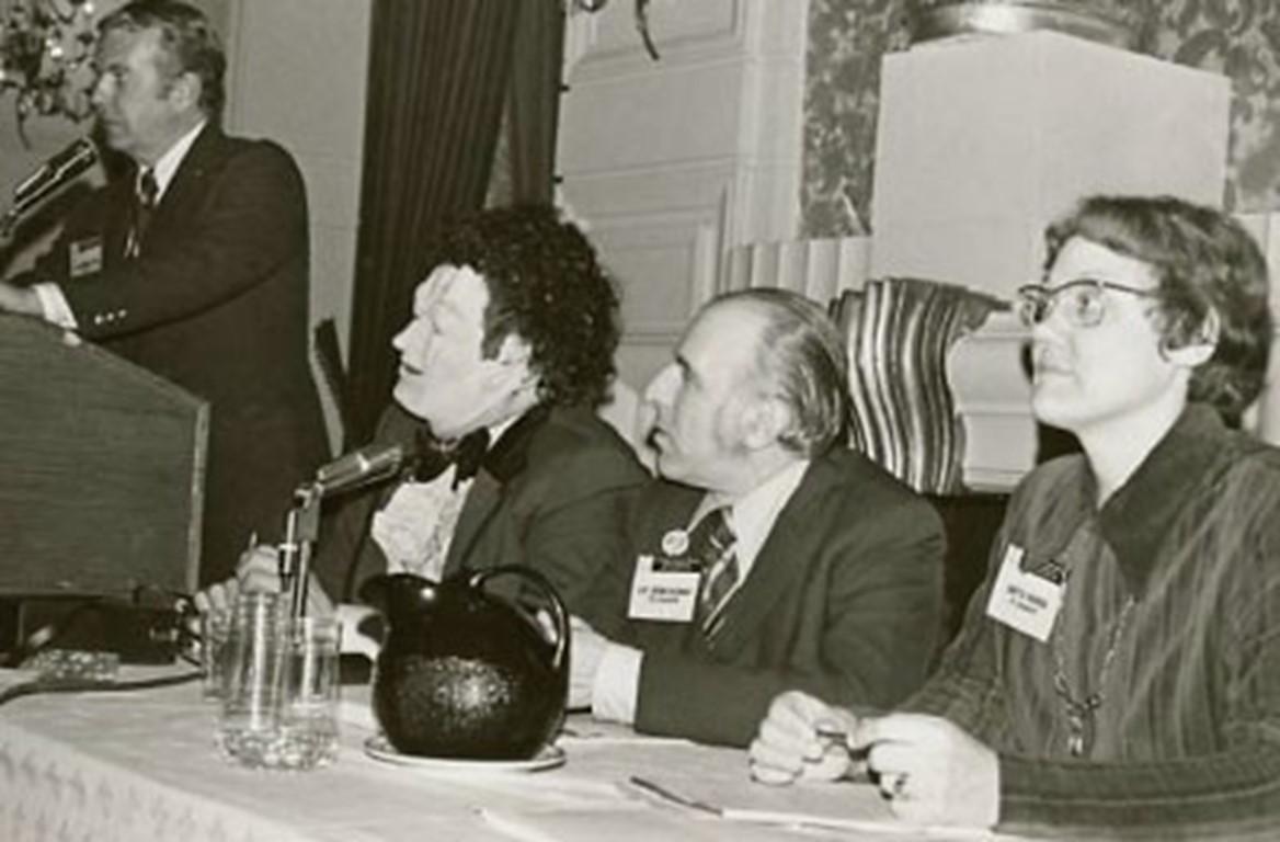 people sitting behind a table with microphones. One person standing at a podium speaking